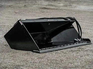 The image shows a black bucket sitting on top of a dirt road. The bucket is upright and empty. The dirt road is winding and dusty, and there are a few trees in the distance. The image is well-lit and in focus.